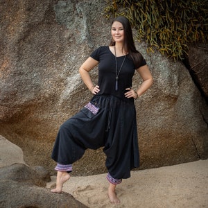 May include: A woman wearing a black t-shirt with a silver necklace and black pants with a colorful patterned trim. She is standing in front of a large rock.