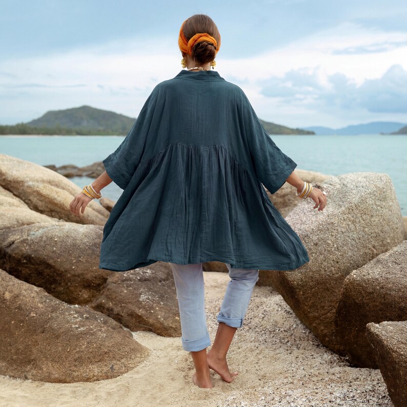 Back view of the Smoky Jade Aria Tunic revealing the full oversized babydoll silhouette and flowing cotton gauze fabric. The wide kimono sleeves and relaxed cut create movement and volume as the model stands on a sandy shoreline with ocean and rocks in the background. The piece highlights breathable comfort and timeless bohemian style.