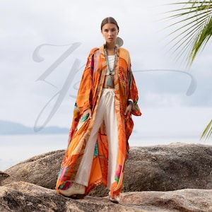May include: A woman wearing an orange, patchwork kimono with a white top and pants. The kimono has a vibrant, colorful design. She is wearing a long necklace with a tassel and large earrings. The background is a beach scene.