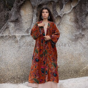 Puede incluir: Una larga bata de terciopelo color óxido con estampado floral. La bata tiene cuello chal y mangas largas. La modelo lleva la bata abierta, mostrando una camiseta blanca debajo.