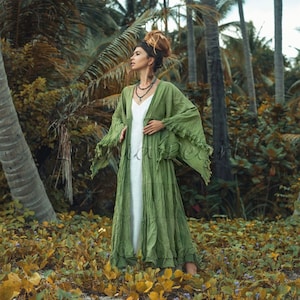May include: A woman wearing a long, green, flowing dress with fringe details. The dress has a white under layer and is open in the front. She is standing in a tropical forest setting.