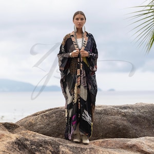May include: A long, flowing kimono-style robe with a patchwork design in shades of black, brown, and pink. The robe is worn over a white top and paired with white boots. The background features a seascape with a cloudy sky.