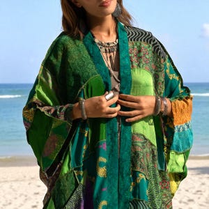 Close-up of the Harper Jade Kimono in Emerald Oasis, revealing intricate patchwork silk details with emerald, teal, saffron, and floral motifs. Styled with silver jewelry and layered over a neutral dress, this eco-friendly bohemian robe reflects artisan craftsmanship. Lightweight and breathable, it’s a one-of-a-kind festival kimono perfect for beach and resort wear.