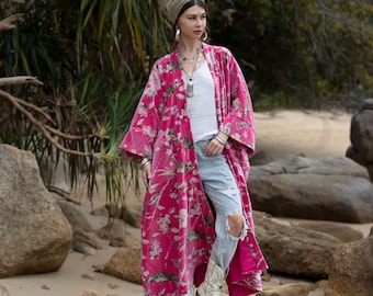 May include: A long pink floral print velvet kimono with a white tank top, blue jeans, and white cowboy boots. The person is wearing a light brown head wrap.