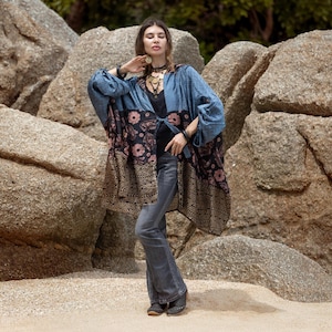 Puede incluir: Una chaqueta estilo kimono de corte holgado con una sección superior de patrón azul, una sección media negra con motivos florales rosas y un dobladillo con patrón en tonos dorados. La prenda se lleva sobre un top negro y vaqueros grises acampanados.