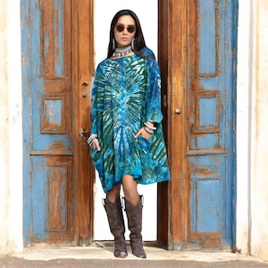 Könnte beinhalten: Eine Frau trägt ein blau-grünes Batik-Kleid mit langen Ärmeln und Taschen, kombiniert mit braunen Cowboy-Stiefeln. Im Hintergrund eine verwitterte blaue Tür und eine braune Tür.