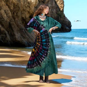 May include: A long, flowing green dress with a tie-dye pattern in shades of purple, blue, yellow, and black. The dress has long sleeves and a button-up front. The model is standing on a sandy beach with the ocean in the background.