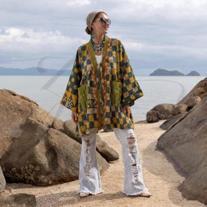 May include: A patchwork kimono-style jacket in shades of yellow, green, and navy blue, featuring a geometric pattern. The jacket has two front pockets and is worn with distressed light-wash jeans. The background shows a seascape with rocks and the ocean.