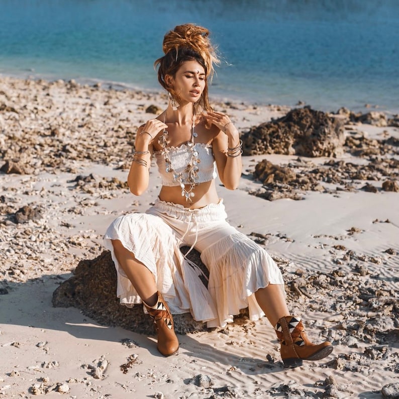 May include: A woman wearing a white crop top and white pants sits on a rock on a beach. She is wearing a silver necklace with a shell design and brown boots with a geometric pattern.