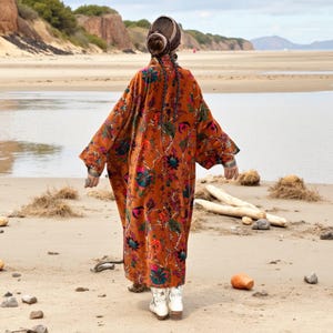 Back view of the Amber Flora Velvet Kimono Robe by ZinniaPearl, showing the sweeping oversized silhouette with jewel-toned botanical patterns across a sienna base. The model walks on sandy terrain near tidal waters, evoking free-spirited boho charm. This plus size velvet duster flows with dramatic presence, featuring wide sleeves, deep pockets, and artisanal detail, ideal for fall fashion, parties, and mindful slow fashion styling.