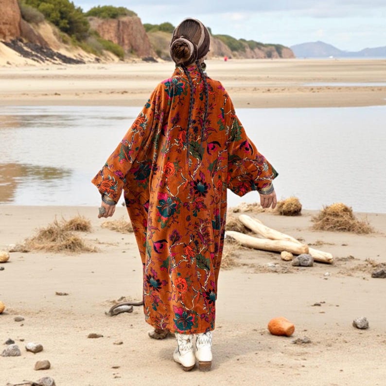 Back view of the Amber Flora Velvet Kimono Robe by ZinniaPearl, showing the sweeping oversized silhouette with jewel-toned botanical patterns across a sienna base. The model walks on sandy terrain near tidal waters, evoking free-spirited boho charm. This plus size velvet duster flows with dramatic presence, featuring wide sleeves, deep pockets, and artisanal detail, ideal for fall fashion, parties, and mindful slow fashion styling.