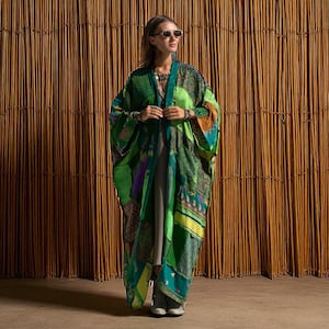 Full-length view of the Harper Jade Kimono in Emerald Oasis, an upcycled vintage silk patchwork robe styled against a natural bamboo backdrop. The jewel-toned green and teal fabrics shimmer in the light, highlighting intricate patterns and one-of-a-kind artistry. This bohemian duster with wide sleeves and flowing length is perfect for festivals, resort wear, and eco-conscious goddess style.