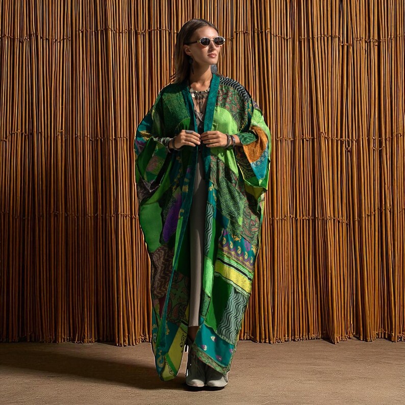 Full-length view of the Harper Jade Kimono in Emerald Oasis, an upcycled vintage silk patchwork robe styled against a natural bamboo backdrop. The jewel-toned green and teal fabrics shimmer in the light, highlighting intricate patterns and one-of-a-kind artistry. This bohemian duster with wide sleeves and flowing length is perfect for festivals, resort wear, and eco-conscious goddess style.