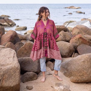 May include: A long-sleeved, button-down tunic featuring a patchwork of magenta, pink, and gold floral patterns with two front pockets, paired with light blue cropped denim jeans. The tunic has a V-neckline and a loose, flowing silhouette.