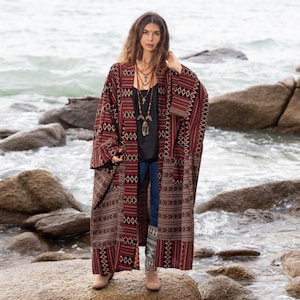 May include: A long, red and black patterned kimono with a geometric design. The kimono is worn over a black top and blue jeans. The person is standing on a rocky beach.