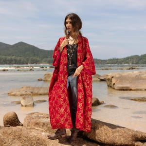 May include: A woman wearing a long, red kimono with gold floral patterns. The kimono has a wide, flowing silhouette and is worn over a black top and blue jeans. The woman is standing on a rocky beach with a blue sky and ocean in the background.