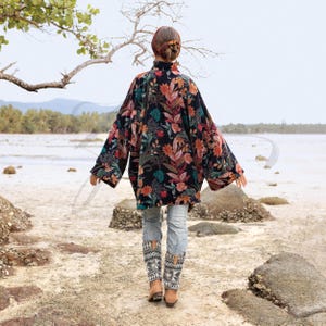 Back view of the Mei Velvet Kimono Duster in Nocturne Flora, showing its wide kimono sleeves and flowing drape as the model walks along tide-washed rocks. The floral motifs in rust, teal, and berry tones bloom across the black velvet canvas. This image evokes serene movement, artisanal texture, and ZinniaPearl’s signature boho-luxury craftsmanship.
