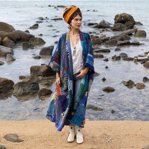 Puede incluir: Una mujer con una chaqueta larga acolchada de patchwork azul y multicolor con una camisa blanca y pantalones vaqueros azules. Está de pie en una playa con rocas y agua en el fondo.