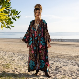 Könnte beinhalten: Langer, offen geschnittener brauner Robe mit farbenfrohen Blumenstickereien in Rosa, Türkis und Gold. Die Robe wird an einem Sandstrand mit dem Meer im Hintergrund getragen. Die Ärmel sind weit und die Robe reicht bis zu den Knöcheln.