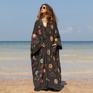 May include: A long, dark grey kimono-style robe featuring colourful embroidered celestial designs, including planets, stars, and clouds. The robe has long sleeves and pockets, and is worn on a sandy beach with the sea in the background.
