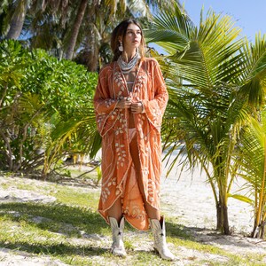 May include: A woman wearing a long, orange embroidered kimono with white floral designs. She is standing in front of palm trees and wearing white cowboy boots.