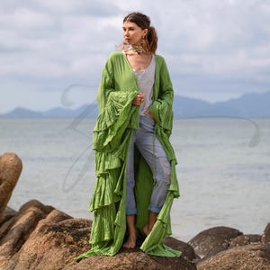 May include: A long, flowing, lime green kimono-style robe with tiered ruffles. The robe has wide sleeves and is worn over a white top and distressed blue jeans. The background features a seascape with a cloudy sky.