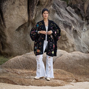 May include: A black kimono jacket, adorned with embroidered planets and stars in various colours, worn with a white top and distressed light-wash jeans. The jacket's design includes geometric shapes. The model is wearing a black headband and hoop earrings.