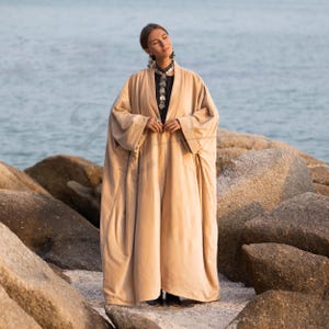 Può includere: Una lunga e fluente veste beige con maniche ampie. La veste è aperta sul davanti, rivelando un top nero e gioielli d'argento. La persona è in piedi su una spiaggia rocciosa con l'oceano sullo sfondo. La veste sembra essere fatta di un tessuto morbido e leggero.
