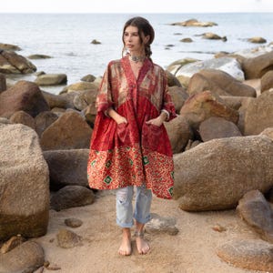 May include: A red and beige patterned kimono-style jacket with pockets. The jacket has a V-neck and is knee-length. The bottom portion of the jacket features a repeating geometric pattern in red, green, and beige. The model is wearing light blue jeans.