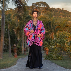 May include: A woman wearing a pink floral embroidered jacket with black trim and a black maxi skirt. She is standing on a path in a tropical garden.