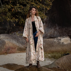 Op de afbeelding: Een vrouw draagt een lange, beige kimono met een bloemenprint. De kimono heeft een wijde, vloeiende silhouet en heeft een delicaat bloemenpatroon in roze, wit en groene tinten. De vrouw staat op een rotsachtig strand, en de achtergrond is een weelderig groen bos.