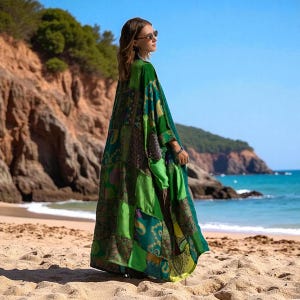 Side view of the Harper Jade Emerald Oasis Kimono, captured against a coastal backdrop with cliffs and bright blue sea. The upcycled vintage silk patchwork design highlights paisley and mandala motifs in rich greens and blues. Lightweight and flowing, this bohemian robe is perfect for resort layering, festivals, or eco-conscious fashion lovers.