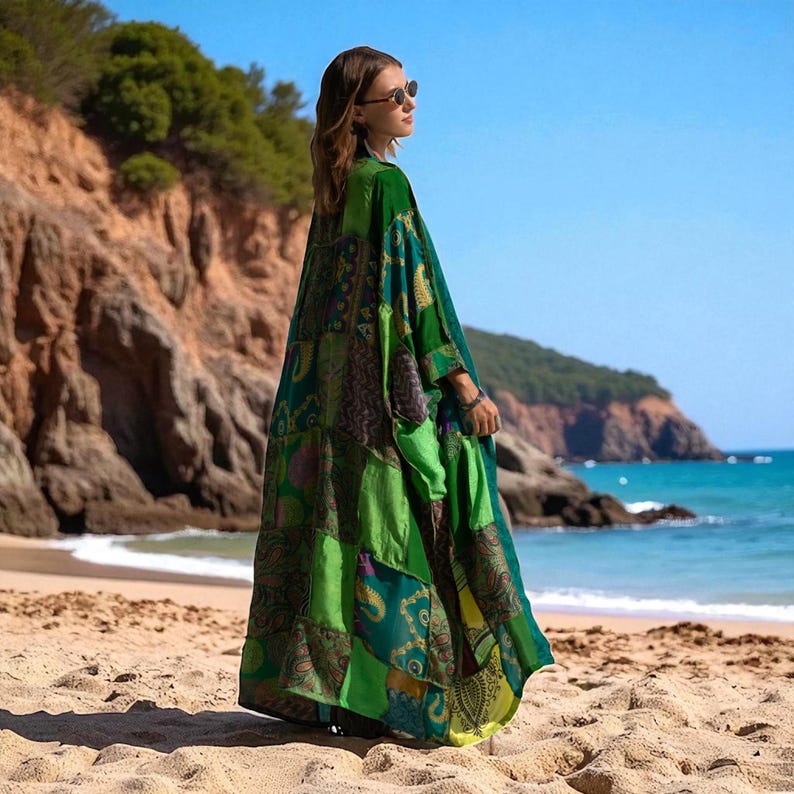 Side view of the Harper Jade Emerald Oasis Kimono, captured against a coastal backdrop with cliffs and bright blue sea. The upcycled vintage silk patchwork design highlights paisley and mandala motifs in rich greens and blues. Lightweight and flowing, this bohemian robe is perfect for resort layering, festivals, or eco-conscious fashion lovers.