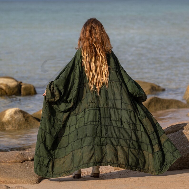 Back view of the Green VEDA kimono showing its dramatic width. The oversized garment flares out, displaying the intricate grid of patchwork seams. The deep moss green canvas contrasts with the dark stitching. A statement piece that bridges the gap between festival wear and architectural fashion.