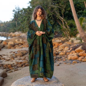 May include: A woman wearing a long, green and blue patchwork robe with a V-neckline. She is standing on a rock with a beach and palm trees in the background.