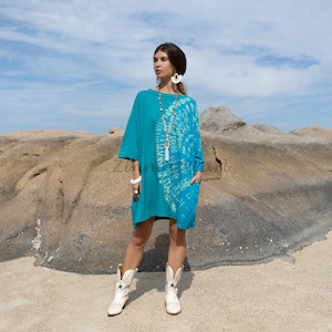 May include: A turquoise blue tie-dye dress with a white and green swirl pattern. The dress has a round neckline and three-quarter sleeves. The model is wearing white cowboy boots.