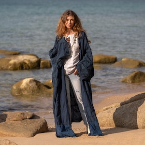 May include: A long, navy blue duster coat with a patchwork design. The coat is worn over a white top and light-wash jeans. The model is standing on a sandy beach with the ocean in the background. The coat has long sleeves and a flowing silhouette.