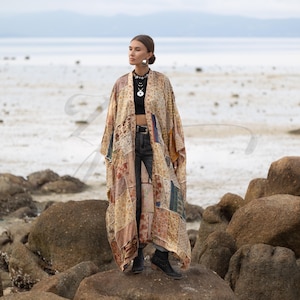 May include: A long patchwork kimono in beige, brown, and blue hues. The kimono is made from various patterned fabric squares. It is worn open, revealing a black top and dark jeans. The background shows a beach and the sea.