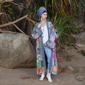 Könnte beinhalten: Ein langer Patchwork-Kimono in Blau-, Grau- und verschiedenen Farbtönen. Der Kimono weist eine Mischung aus gemusterten Stoffen und einen kontrastierenden Besatz auf. Die Person trägt ein weißes Oberteil, blaue Jeans und weiße Stiefel. Ein blaues Haarband und Schmuck vervollständigen den Look.