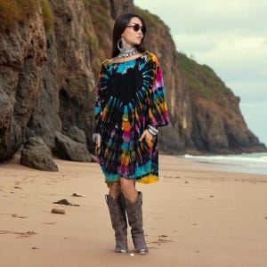 May include: A tie-dye dress with a black heart design, featuring a mix of vibrant colors like blue, yellow, and pink. The dress has long sleeves and pockets. The person is wearing silver jewelry and brown cowboy boots. The background is a beach with cliffs.