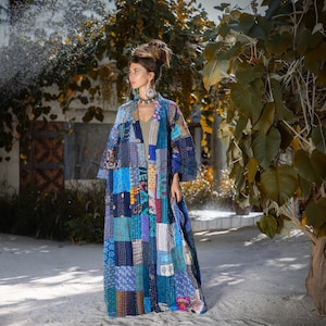May include: A woman wearing a long, blue and multi-coloured patchwork quilted jacket. The jacket features a variety of patterns and textures, including floral, geometric, and paisley designs. The woman is standing in front of a green leafy bush.