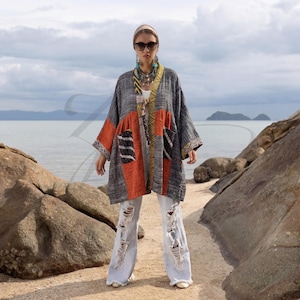 May include: A gray, orange, and black patterned kimono-style jacket with long sleeves and pockets. The jacket is worn with light blue ripped jeans. The background shows a beach and ocean.