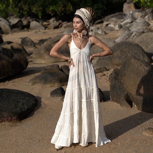 A boho beach wedding dress handcrafted from breathable raw cotton, the Savita Gown is a dreamy choice for elopements, barefoot ceremonies, and destination nuptials. Featuring a plunging neckline, adjustable tie straps, and smocked back, this tiered maxi dress flows with lightweight grace and earthy elegance. Perfect for brides seeking a minimalist yet soulful bridal look with natural fibers, ethical craftsmanship, and plus-size inclusivity.