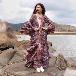 Può includere: Una donna indossa un lungo abito a fiori viola e rosso con balze. L'abito ha maniche lunghe con polsini a campana e una profonda scollatura a V. È in piedi su una spiaggia rocciosa con l'oceano sullo sfondo.