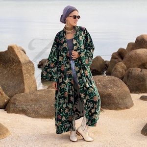 May include: A long, dark green velvet kimono with a floral pattern in shades of orange, pink, and yellow. The kimono is open in the front and has wide sleeves. The garment is worn over a gray top and jeans, paired with white boots.