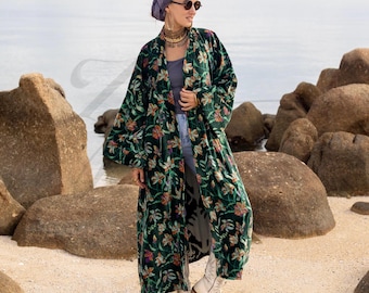 May include: A long, dark green velvet kimono with a floral pattern in orange, pink, and yellow. The kimono is open-fronted and has wide sleeves. The garment is worn over a grey top and jeans, paired with white boots.
