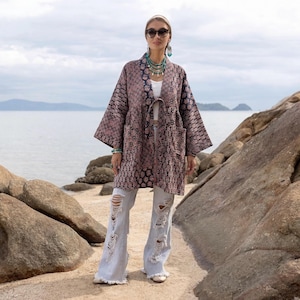 May include: A long-sleeved, open-front jacket featuring a dark blue and pink intricate floral pattern, two front patch pockets, and a tie closure at the waist. The jacket is worn over a white top paired with light blue distressed flare jeans.
