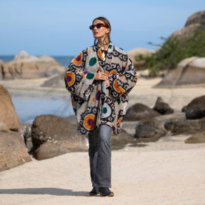 May include: A patterned, oversized coat in shades of gray, black, orange, purple, and green. The coat features a floral design and is worn with dark gray jeans. The background shows a beach with rocks and the ocean.