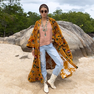Könnte beinhalten: Senfgelber Kimono mit floralem Muster und weiß-gelbem Futter. Er wird über einem pfirsichfarbenen Oberteil und hellblauen zerrissenen Jeans getragen. Weiße Cowboy-Stiefel vervollständigen den Look. Accessoires: braunes Stirnband, Sonnenbrille und mehrreihige Halsketten.