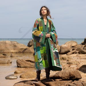 May include: A long green patchwork quilted robe with a tie closure. The robe is made of various fabrics in shades of green, blue, and brown. The model is standing on a rocky beach.
